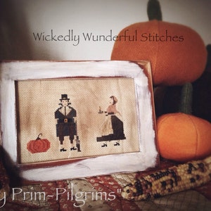 May include: A white framed cross-stitch embroidery depicting two figures in pilgrim clothing, a man and a woman, standing next to a small pumpkin. The embroidery is titled "Thy Prim-Pilgrims" and is surrounded by fall-themed decorations, including pumpkins and corn.