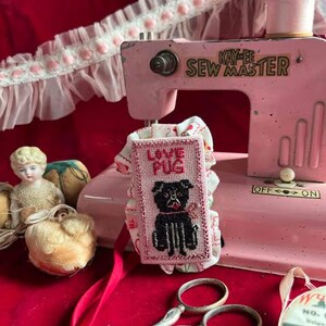 May include: A pink vintage sewing machine with a white and pink fabric with the words "Love Pug" embroidered on it. The fabric is attached to the sewing machine with a pink ribbon. There is a pair of silver scissors and a spool of white thread on a red surface.
