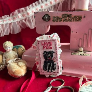 May include: A pink vintage sewing machine with a white and pink fabric featuring the words "Love Pug" embroidered on it. The fabric is attached to the sewing machine with a pink ribbon. There are a pair of silver scissors and a spool of white thread on a red surface.