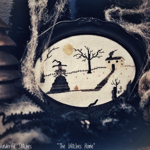 May include: A framed cross-stitch design of a witch's home with a witch standing in front of it. The design is in black and white with a few orange accents. The frame is black and the background is a dark, blurry image of a skull and spiderwebs.