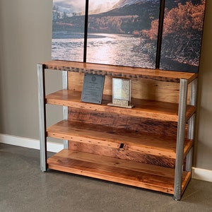 May include: A rustic wooden bookshelf with three shelves and a metal frame. The wood has a natural grain and a warm, reddish-brown hue. The shelves are supported by vertical metal posts. A landscape painting hangs above the bookshelf.