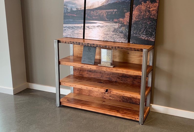 May include: A rustic wooden bookshelf with three shelves and a metal frame. The wood has a natural grain and a warm, reddish-brown hue. The shelves are supported by vertical metal posts. A landscape painting hangs above the bookshelf.