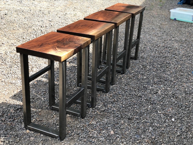 以下が含まれることがあります： 黒い金属製のフレームが付いた木製のバーチェアが4つあります。チェアは一列に並んでおり、各チェアの座面は視聴者の方を向いています。チェアは自然な木で作られており、豊かな茶色をしています。金属製のフレームは頑丈で、黒い仕上げになっています。