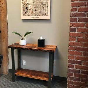 May include: A wooden console table with a black metal frame. The table has a single shelf and a top surface with a natural wood grain. A small potted plant and a black tissue box are on the table.