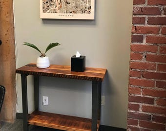 May include: A wooden console table with a black metal frame. The table has a single shelf and a top surface with a natural wood grain. A small potted plant and a black tissue box are on the table.