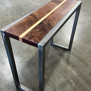 Pu&ograve; includere: Un moderno tavolino consolle con piano in legno scuro con una striscia di legno chiaro al centro. Il piano &egrave; incorniciato da un bordo in metallo argentato e sostenuto da gambe in metallo argentato.