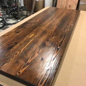 May include: A close-up of a long, rectangular wooden table top with a dark brown stain. The wood grain is visible and has a rustic, reclaimed look.