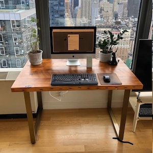 May include: A wooden desk with gold metal legs. The desk has a computer, keyboard, mouse, and two potted plants on it. The desk is in front of a window with a view of a city skyline.