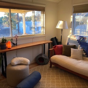 May include: A living room with a futon couch, a wooden table, a lamp, and a window with a view of trees. The futon is covered in a patchwork quilt and has several pillows. The table is made of wood and has a metal frame. The lamp is on a table and has a white shade. The window is open and shows a view of trees outside.