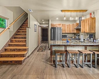 May include: An open-plan interior featuring a wooden staircase with dark stained treads and a matching handrail. The kitchen area includes light wood cabinets, a granite worktop island, and four stools with metal frames and wooden seats.