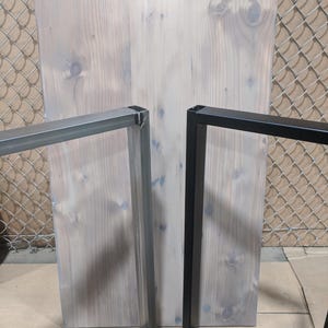 May include: Two metal table legs, one black and one gray, are shown against a light wood background. The legs are made of square metal tubing and have a simple, industrial design.