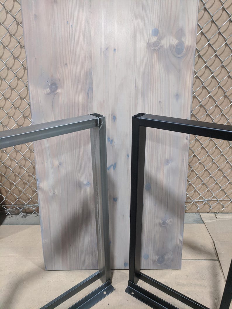 May include: Two metal table legs, one black and one gray, are shown against a light wood background. The legs are made of square metal tubing and have a simple, industrial design.