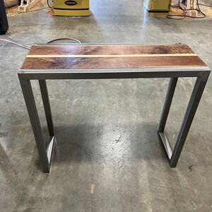 Pu&ograve; includere: Tavolino rettangolare con piano in legno scuro e una striscia di legno chiaro al centro. Il tavolo ha una struttura in metallo con gambe angolate. Adatto come scrivania o tavolino, lungo circa 100 cm.