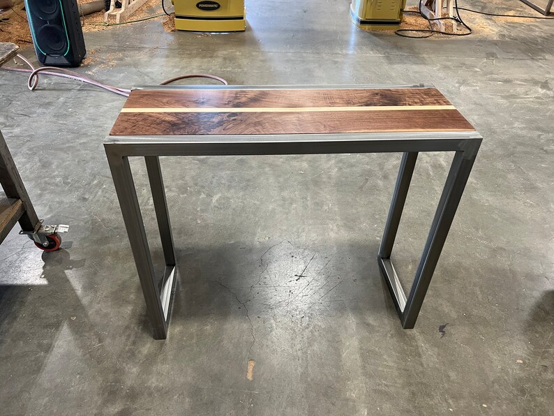 Pu&ograve; includere: Tavolino rettangolare con piano in legno scuro e una striscia di legno chiaro al centro. Il tavolo ha una struttura in metallo con gambe angolate. Adatto come scrivania o tavolino, lungo circa 100 cm.