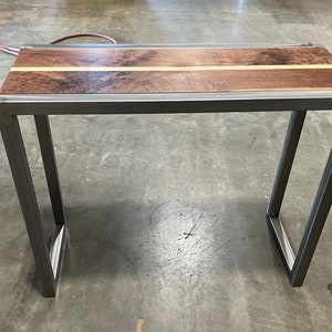 Pu&ograve; includere: Un tavolo consolle artigianale con piano in legno di noce scuro e una striscia decorativa in legno chiaro. Il tavolo ha una robusta struttura in metallo grigio scuro. Il tavolo &egrave; lungo circa 122 cm, largo 30 cm e alto 76 cm.