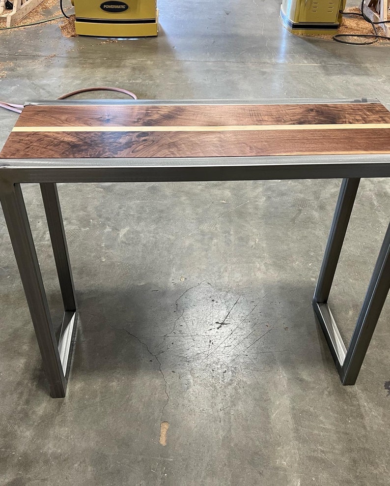 Pu&ograve; includere: Un tavolo consolle artigianale con piano in legno di noce scuro e una striscia decorativa in legno chiaro. Il tavolo ha una robusta struttura in metallo grigio scuro. Il tavolo &egrave; lungo circa 122 cm, largo 30 cm e alto 76 cm.