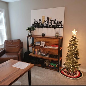 May include: A living room scene featuring a wooden bookshelf with metal supports, filled with books and decor. Above the shelf is a large canvas painting of a black and gold cityscape. A small Christmas tree with lights and a star topper stands nearby.
