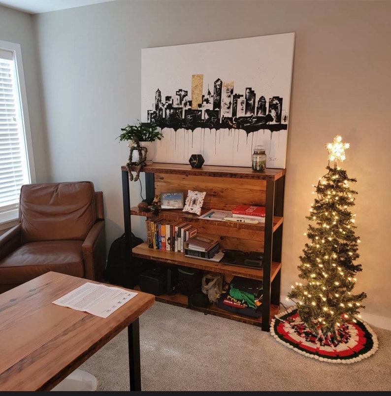 May include: A living room scene featuring a wooden bookshelf with metal supports, filled with books and decor. Above the shelf is a large canvas painting of a black and gold cityscape. A small Christmas tree with lights and a star topper stands nearby.