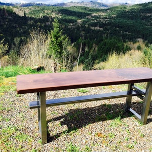 May include: A wooden bench with a dark brown top and a metal frame. The bench is sitting on a gravel surface with green grass in the background.