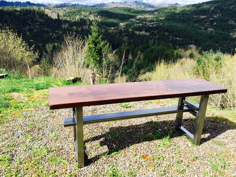 May include: A wooden bench with a dark brown top and a metal frame. The bench is sitting on a gravel surface with green grass in the background.