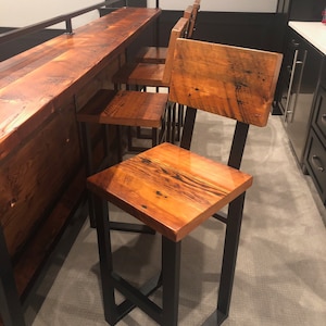 May include: Four wooden bar stools with black metal frames. The stools have a natural wood finish and are arranged in a row. The stools are in front of a wooden bar counter.