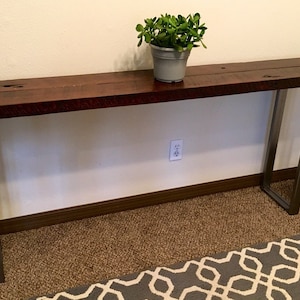 May include: A long, dark wood console table with metal legs. The table is sitting on a brown carpet with a gray and white patterned rug in front of it. A small potted plant sits on the table.