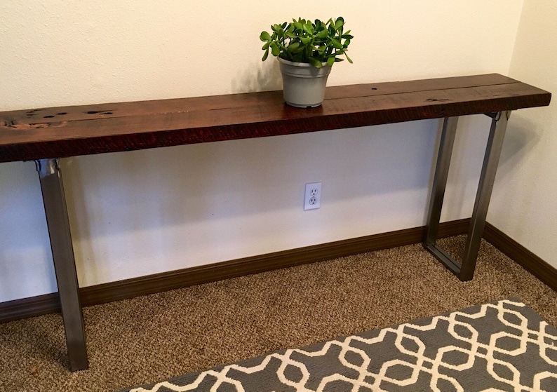 May include: A long, dark wood console table with metal legs. The table is sitting on a brown carpet with a gray and white patterned rug in front of it. A small potted plant sits on the table.