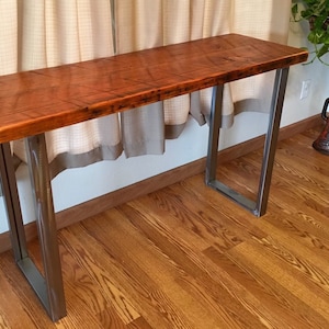 May include: A rustic wooden table top with a natural wood finish sits on a metal frame with a black powder coat finish. The table is in a room with hardwood floors.