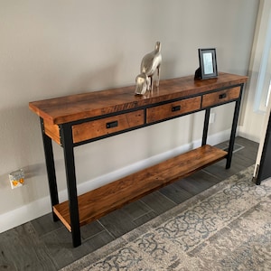May include: A long, rustic wooden console table with three drawers and a lower shelf. The table has a black metal frame and is sitting on a tiled floor.