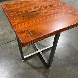 May include: A small, square wooden table with a dark reddish-brown finish. The table has a simple, modern design with a metal base that is painted black.
