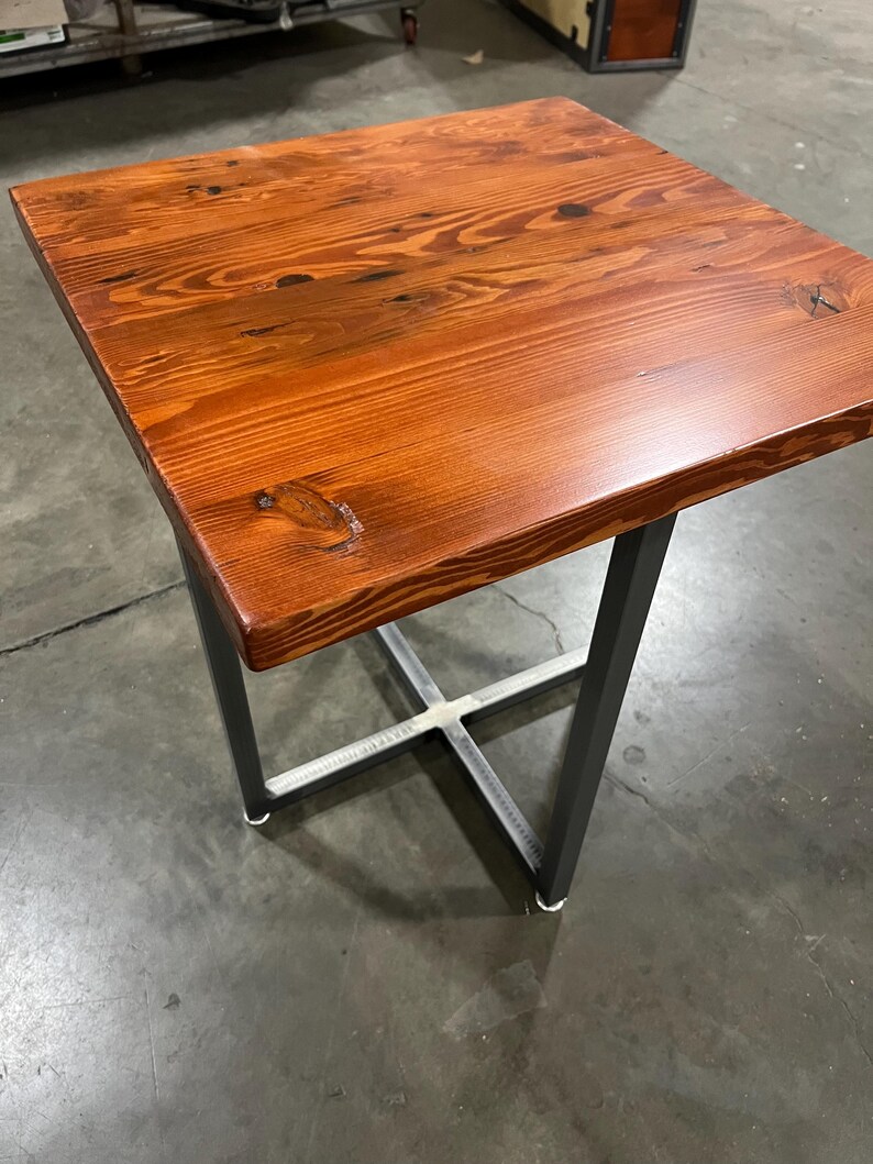 May include: A small, square wooden table with a dark reddish-brown finish. The table has a simple, modern design with a metal base that is painted black.