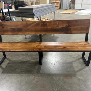 May include: A wooden bench with a backrest and black metal legs. The bench has a natural wood finish and is made of reclaimed wood.
