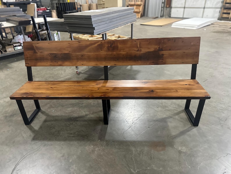 May include: A wooden bench with a backrest and black metal legs. The bench has a natural wood finish and is made of reclaimed wood.