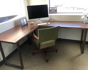 Puede incluir: Un escritorio en forma de L con una superficie de madera de veta marrón oscuro y patas de metal negro. Una silla de oficina tapizada en verde con marco de madera y ruedas está colocada frente al escritorio, que sostiene un monitor, teclado y ratón.