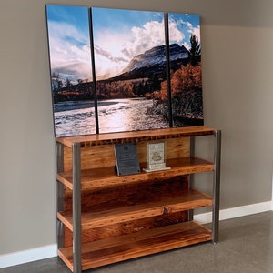 May include: A three-panel landscape print of a mountain and river scene hangs above a wooden bookshelf with metal supports. The bookshelf has three shelves and two plaques on the top shelf. The artwork features a river, trees, and a mountain under a cloudy sky.