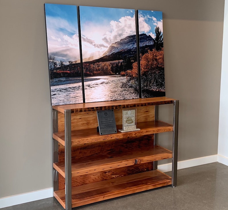 May include: A three-panel landscape print of a mountain and river scene hangs above a wooden bookshelf with metal supports. The bookshelf has three shelves and two plaques on the top shelf. The artwork features a river, trees, and a mountain under a cloudy sky.