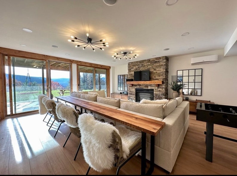 May include: A large, light-colored sectional sofa with multiple pillows sits in front of a stone fireplace with a wood mantel. A long wooden table with black metal legs extends from the sofa, with white furry chairs on either side. The room has a large window with a view of a wooded area and a patio.