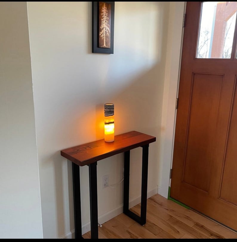 May include: A wooden table with black metal legs and a white and black striped lamp on top. The table is in a room with a wooden door and white walls.