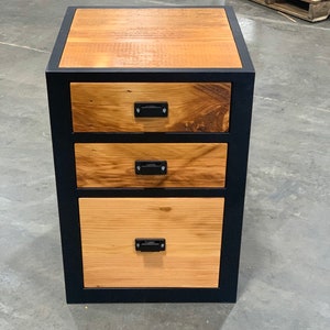 May include: A black metal framed wooden file cabinet with three drawers. The drawers have black metal handles and are made of wood with a natural finish.