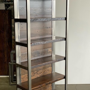 May include: A tall, rustic bookshelf with six shelves. The shelves are made of dark stained wood, and the frame is constructed of silver metal. The back of the bookshelf is made of light-colored wood planks. The bookshelf is designed for storage and display.