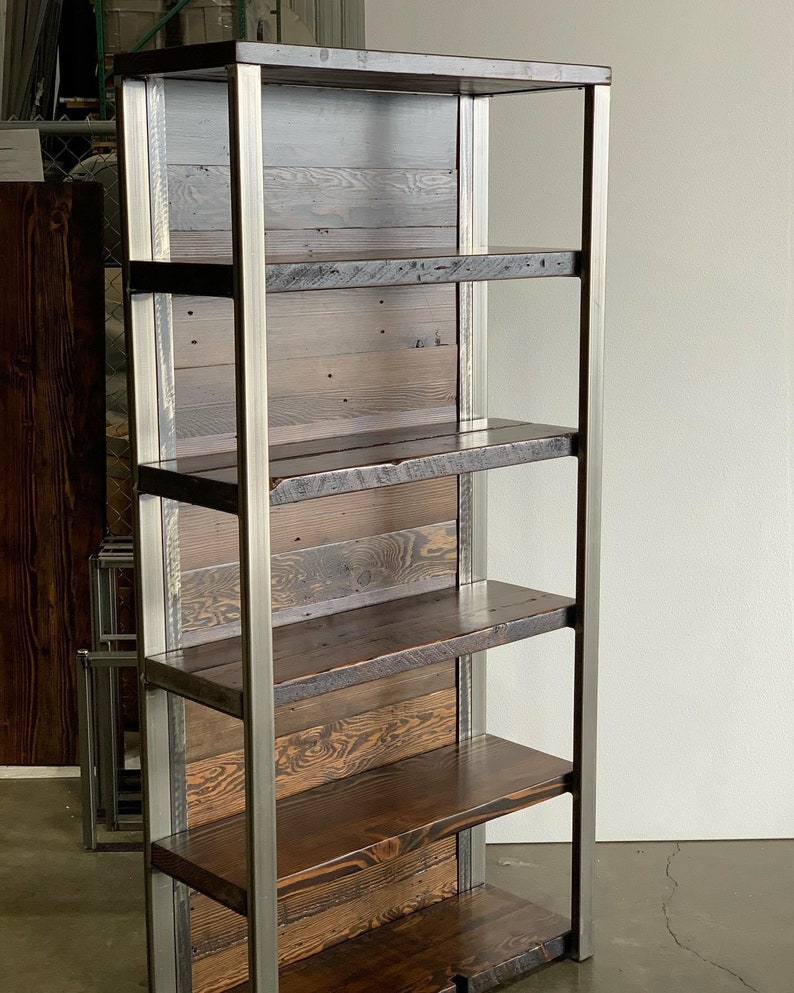 May include: A tall, rustic bookshelf with six shelves. The shelves are made of dark stained wood, and the frame is constructed of silver metal. The back of the bookshelf is made of light-colored wood planks. The bookshelf is designed for storage and display.