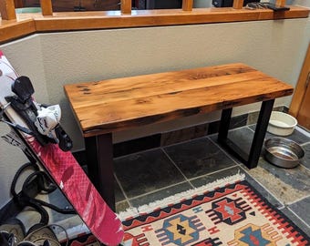 May include: A rectangular bench featuring a natural wood grain top with a smooth finish and four sturdy black metal legs. The bench is positioned horizontally, showcasing a clear wood texture with visible knots and dark brown tones throughout the surface.