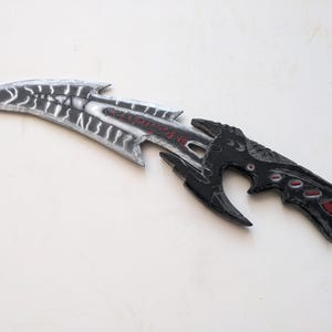 May include: A black and silver fantasy-style dagger with red accents. The blade is curved and has a serrated edge. The handle is black and has a skull-like design.