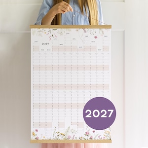 以下が含まれることがあります： 1月から12月までの月が記載された縦型グリッドレイアウトの2027年壁掛けカレンダー。上下に繊細な花のイラストが描かれており、吊り下げ用の紐が付いた明るい木製のフレームで固定されています。