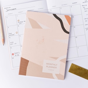 Op de afbeelding: Een maandplanner met een abstract ontwerp in beige, perzik en zwart. De planner is open en toont een septemberkalender. Een potlood en een messing liniaal zijn ook zichtbaar.