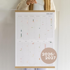 Könnte beinhalten: Ein großer Wandkalender für 2026-2027, gehalten von einem Holzrahmen. Der Kalender hat ein minimalistisches Design mit monatlichen Gittern und farbcodierten Markierungen. Die Monate sind auf Englisch und Deutsch beschriftet. Eine kreisförmige Grafik zeigt "2026/2027".