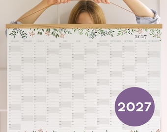 May include: A 2027 wall calendar featuring a horizontal grid layout with twelve months from January to December. The paper is held by light wood magnetic rails at the top and bottom. The design includes botanical illustrations of green leaves and red berries.