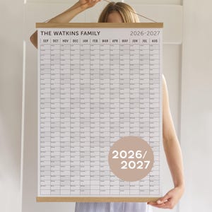 May include: A large wall calendar for 2026-2027, held by a person. The calendar has a white grid layout, wooden frame, and the text "THE WATKINS FAMILY" at the top. The years 2026 and 2027 are also displayed.