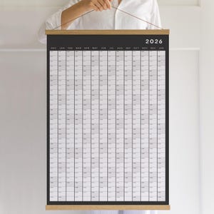 May include: A large, rectangular 2026 year calendar with a black background and a grid of small squares for each day. The calendar is held by a wooden bar at the top and bottom. The months are listed across the top, with the year 2026 in white text.