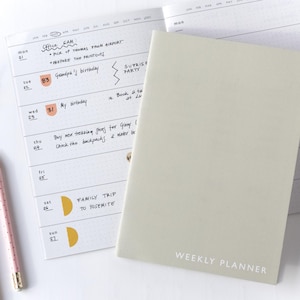 Op de afbeelding: Een weekplanner met een lichtgrijze kaft en de woorden "WEEKLY PLANNER" op de voorkant. De open planner toont een wekelijkse lay-out met handgeschreven notities en data. Een roze en witte potlood is zichtbaar.